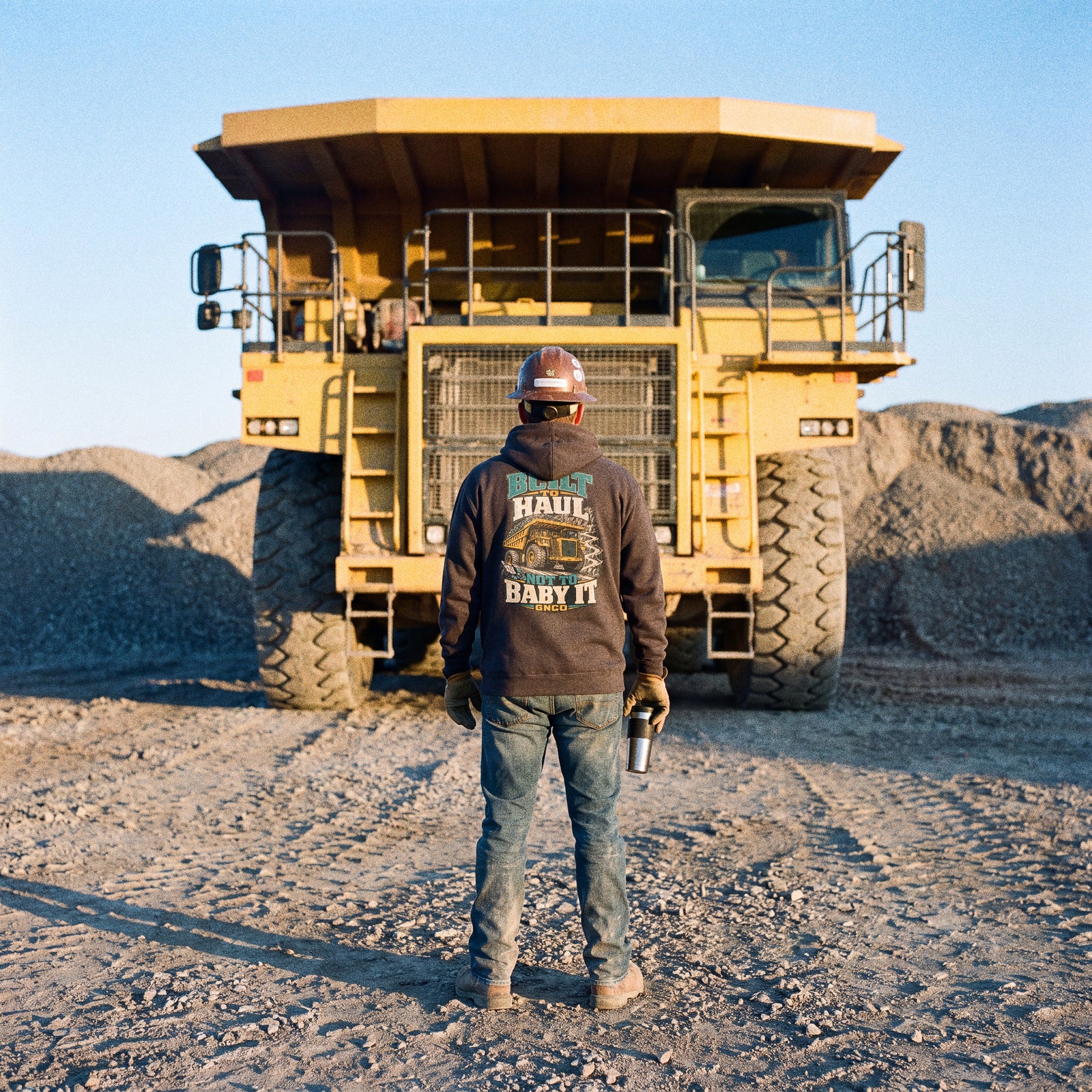 Operator in a BUILT TO HAUL hoodie facing a rigid mining haul truck at first light